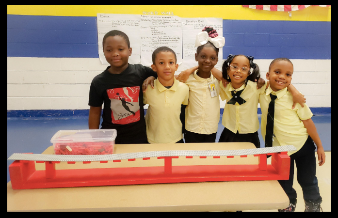  2nd grade students with their arms around each other, standing behind their finished LEGO bridge